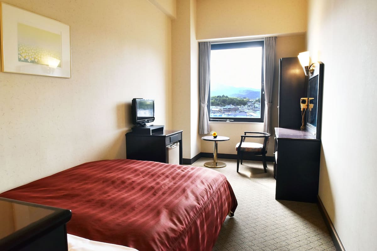 Ett hotellrum med en säng, ett skrivbord med en dator, en stol, ett litet bord med en vas och ett fönster med utsikt över stadsbilden.