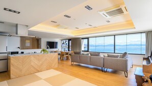 A modern hotel room with a kitchen, a sofa set, a piano, and a large window offering a view of the sea.