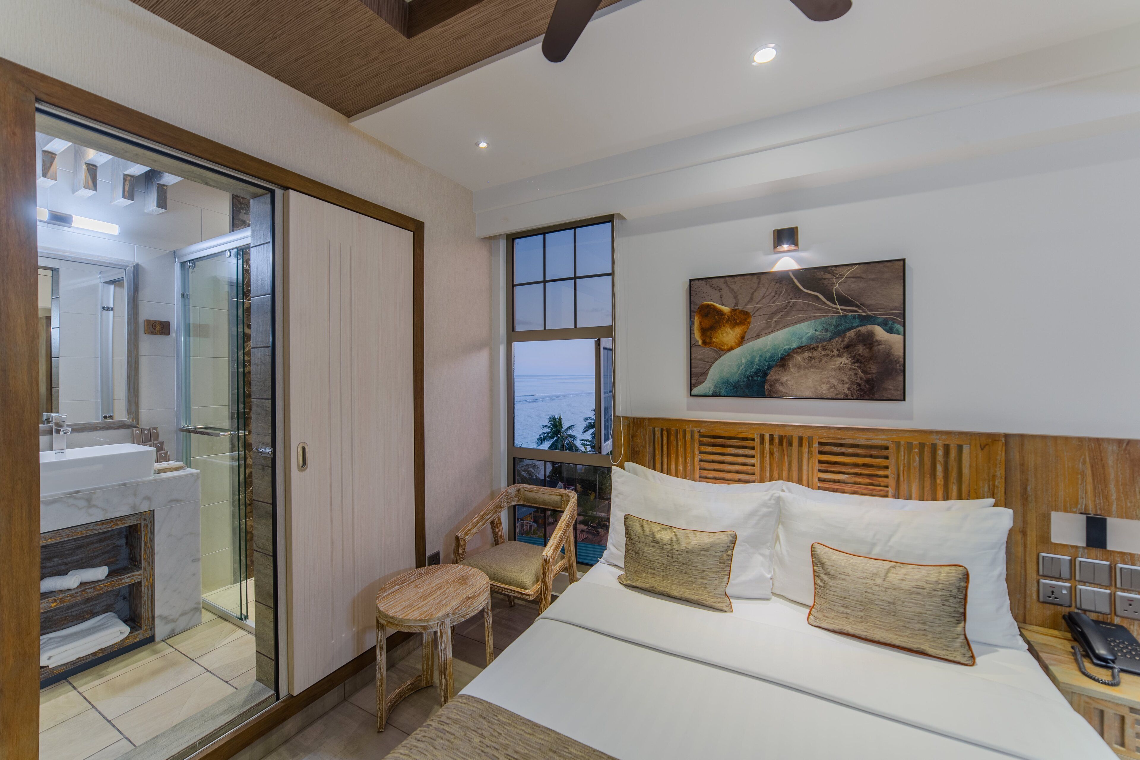 A hotel room with a bed, a chair, a desk with a phone, a painting on the wall, and a view of the ocean through a window.