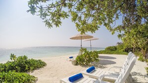 Ein Strandloungebereich mit weißen Liegestühlen und blauen Kissen, einem beige farbenen Sonnenschirm und Blick aufs Meer.