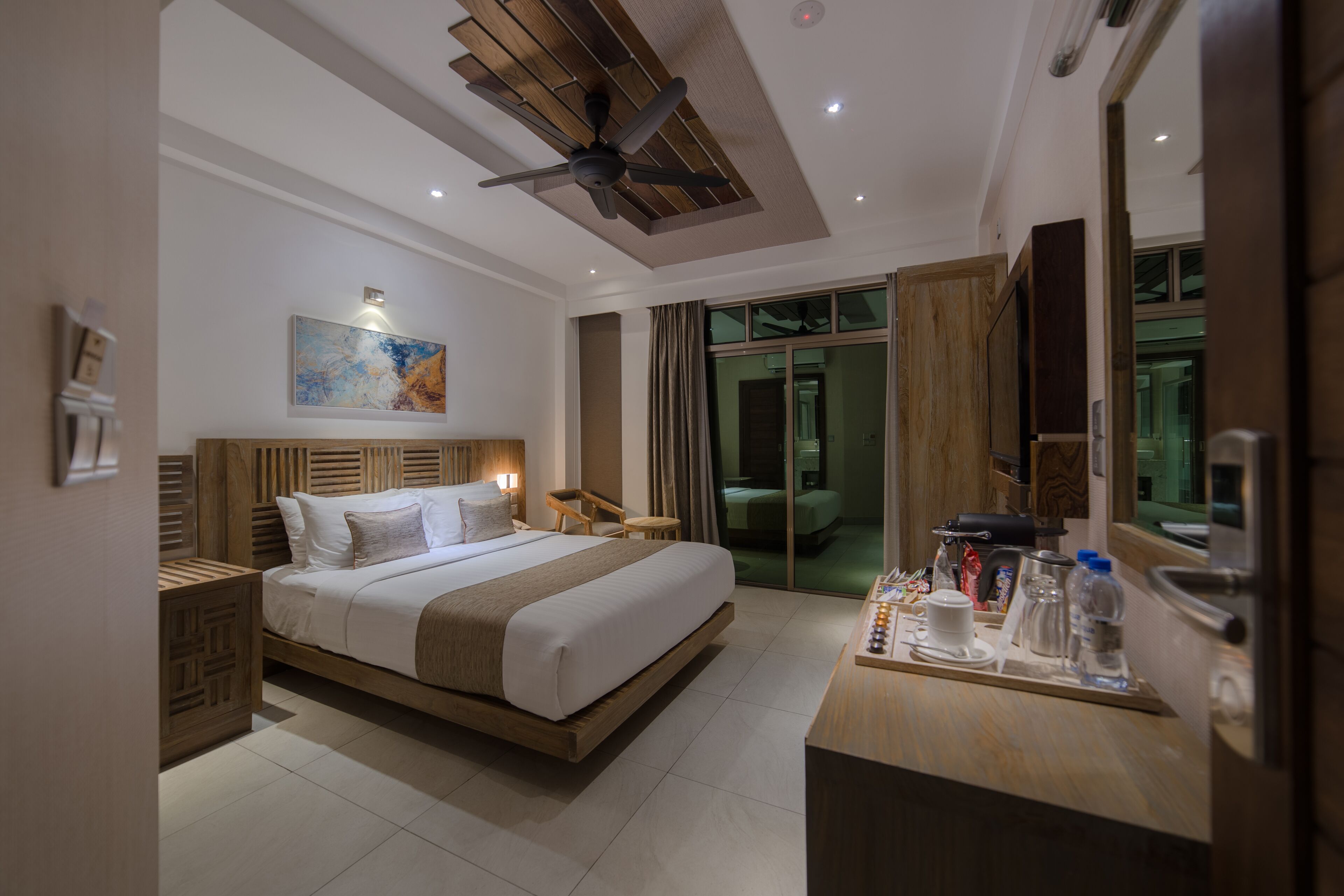 A modern hotel room with a large bed, a wooden headboard, a ceiling fan, a bedside table with a lamp, a small table with a tray of breakfast items, and a large mirror.