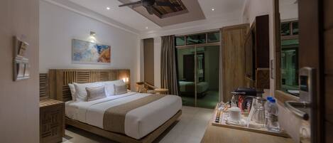 A modern hotel room with a large bed, a wooden headboard, a ceiling fan, a bedside table with a lamp, a small table with a tray of breakfast items, and a large mirror.
