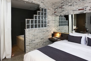 A modern hotel room with a brick wall, a large bed, a nightstand with a lamp, and a bathroom visible through a glass door. - S& Hotel (Daejeon)