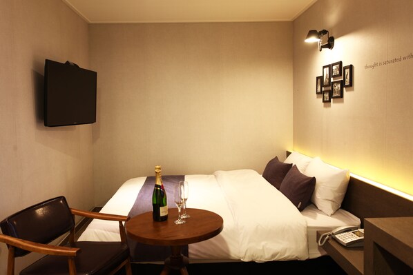 A hotel room with a bed, a bedside table with a bottle and glasses, a chair, a wall-mounted TV, and a wall with framed pictures. - S& Hotel (Daejeon)