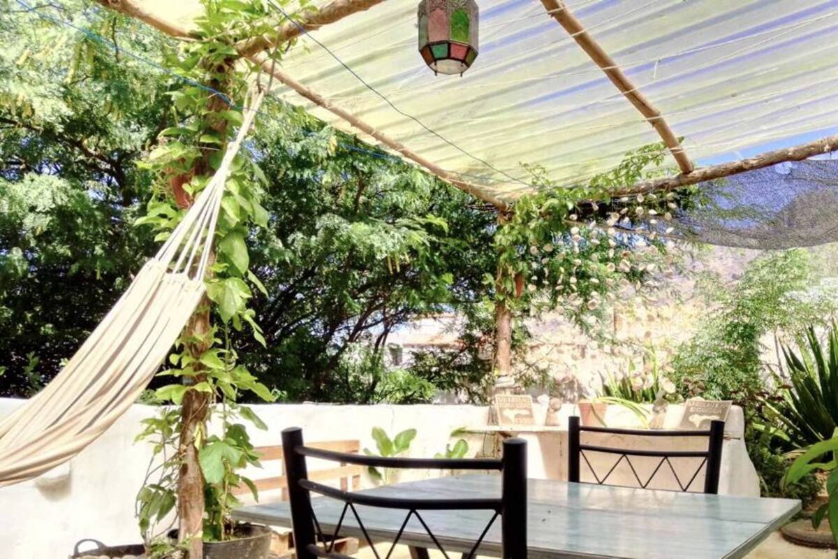 A covered outdoor seating area with a table and chairs, surrounded by greenery and a hammock.