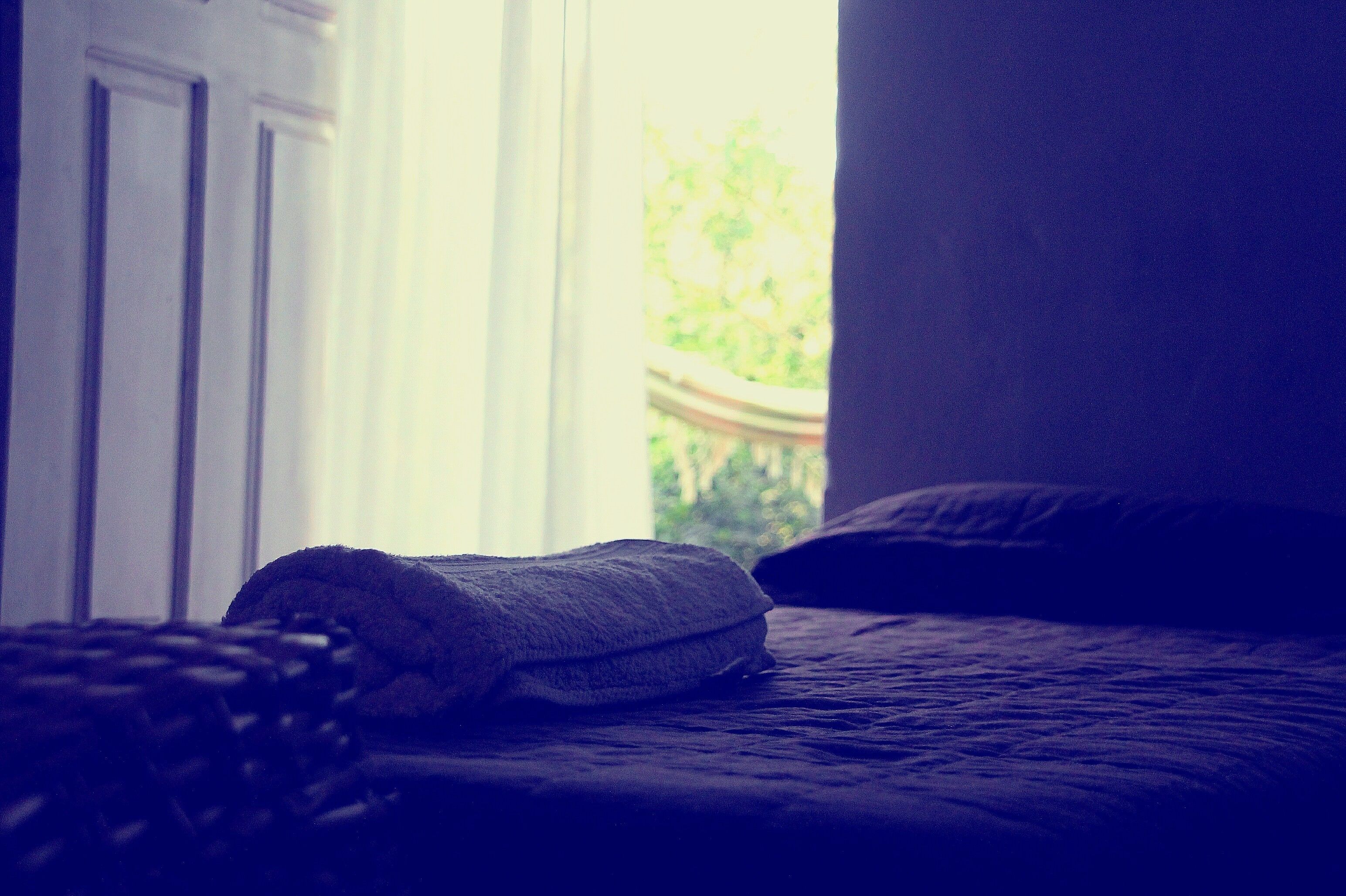 A bed with a folded blanket and pillows, a window with a view of greenery, and a door.