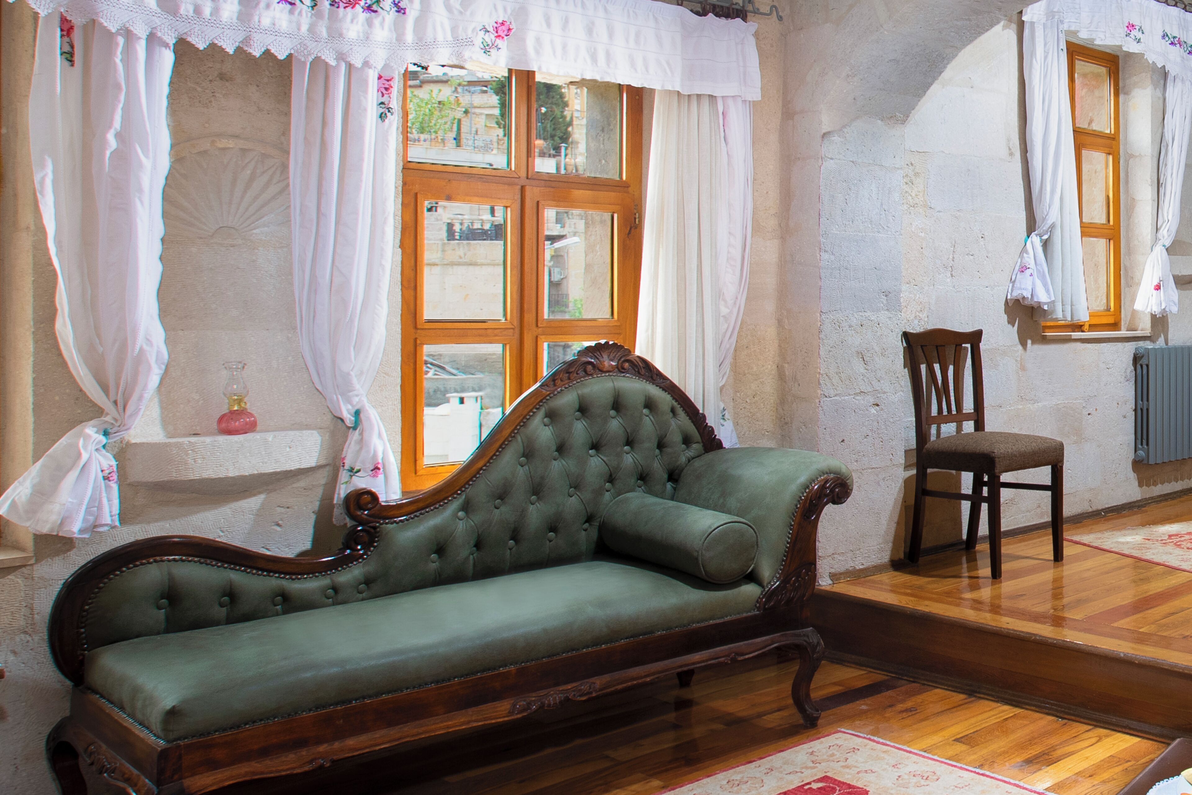 A vintage green sofa with a wooden frame, a wooden chair, and a window with white curtains.
