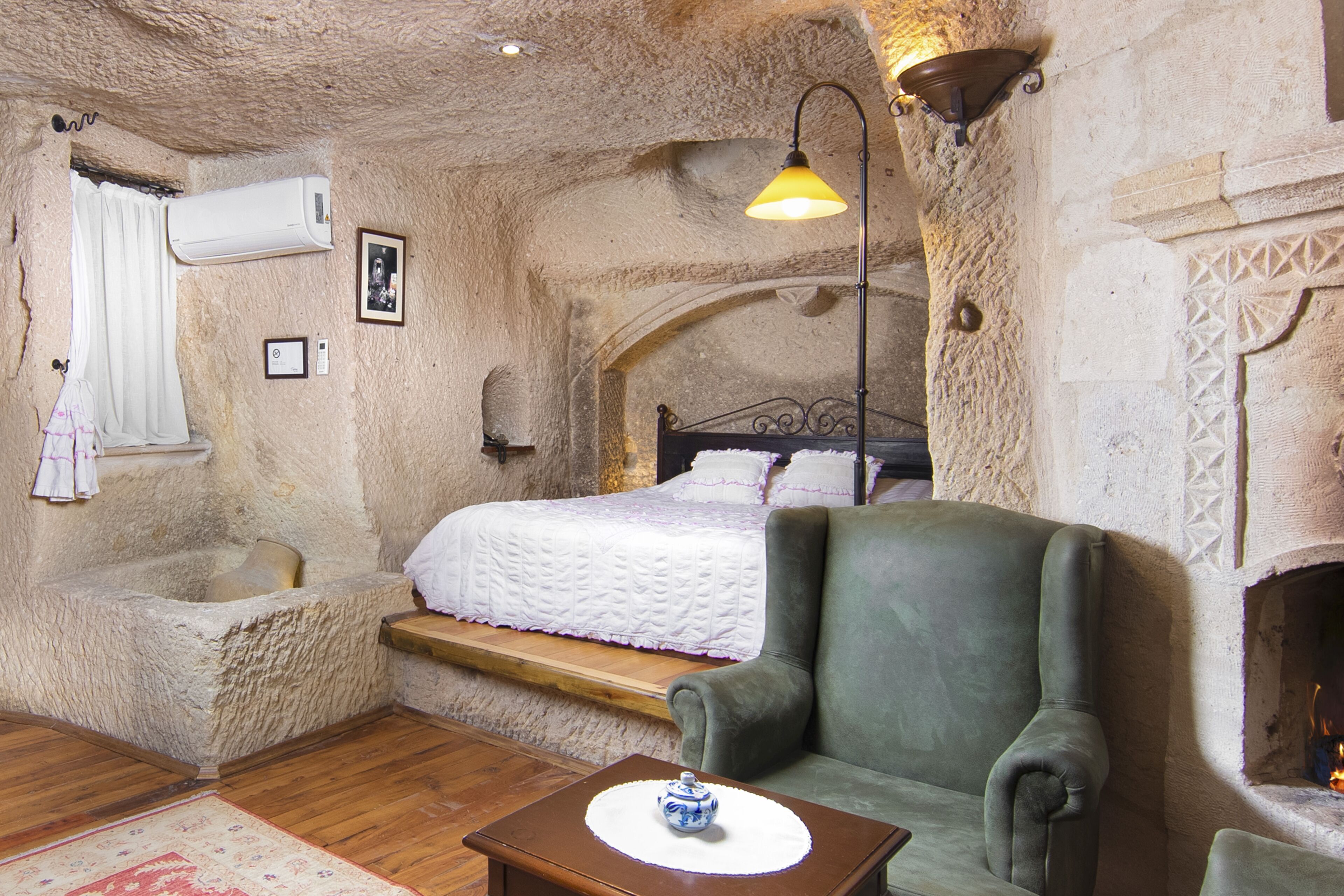 A bedroom with a bed, a green armchair, a bedside table, a fireplace, and a stone wall.
