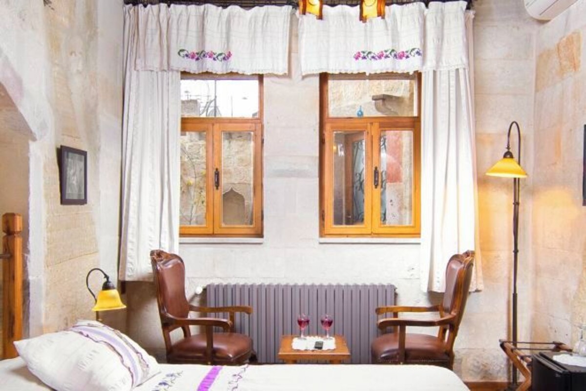 Una habitación con dos ventanas de madera, una cama con colcha blanca, dos sillones de cuero, una mesita con dos copas de vino tinto y una lámpara de pie.