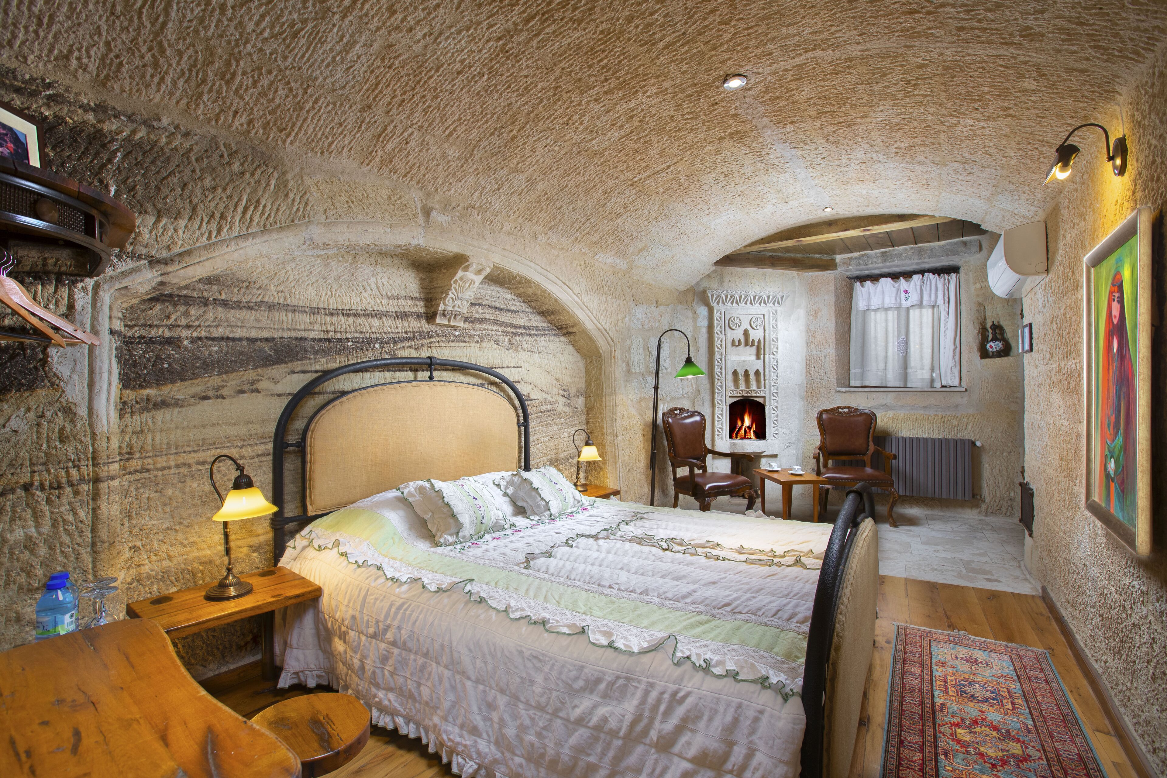 A rustic room with a stone ceiling, a bed with a wooden headboard, a small table with a lamp, and a chair.