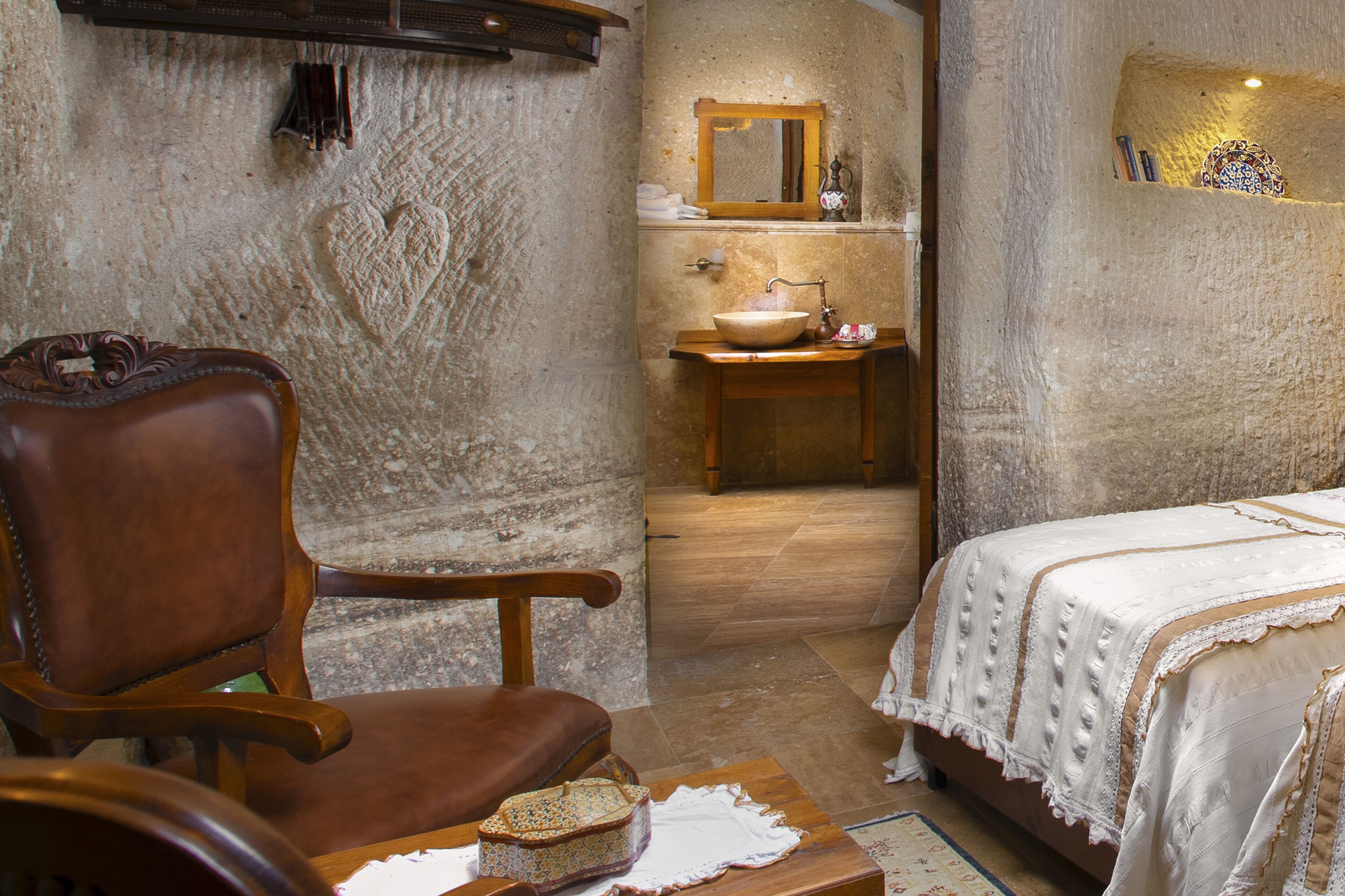 A room with a wooden chair, a bed with a white bedspread, a wooden nightstand, a small table with a bowl, and a mirror above the sink.