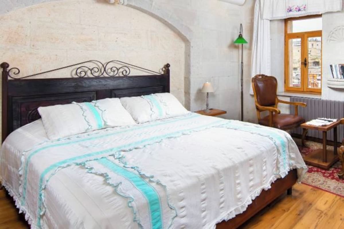 Un dormitorio con cama de madera, ropa de cama blanca, una lámpara de pie verde, una silla marrón y una mesita.