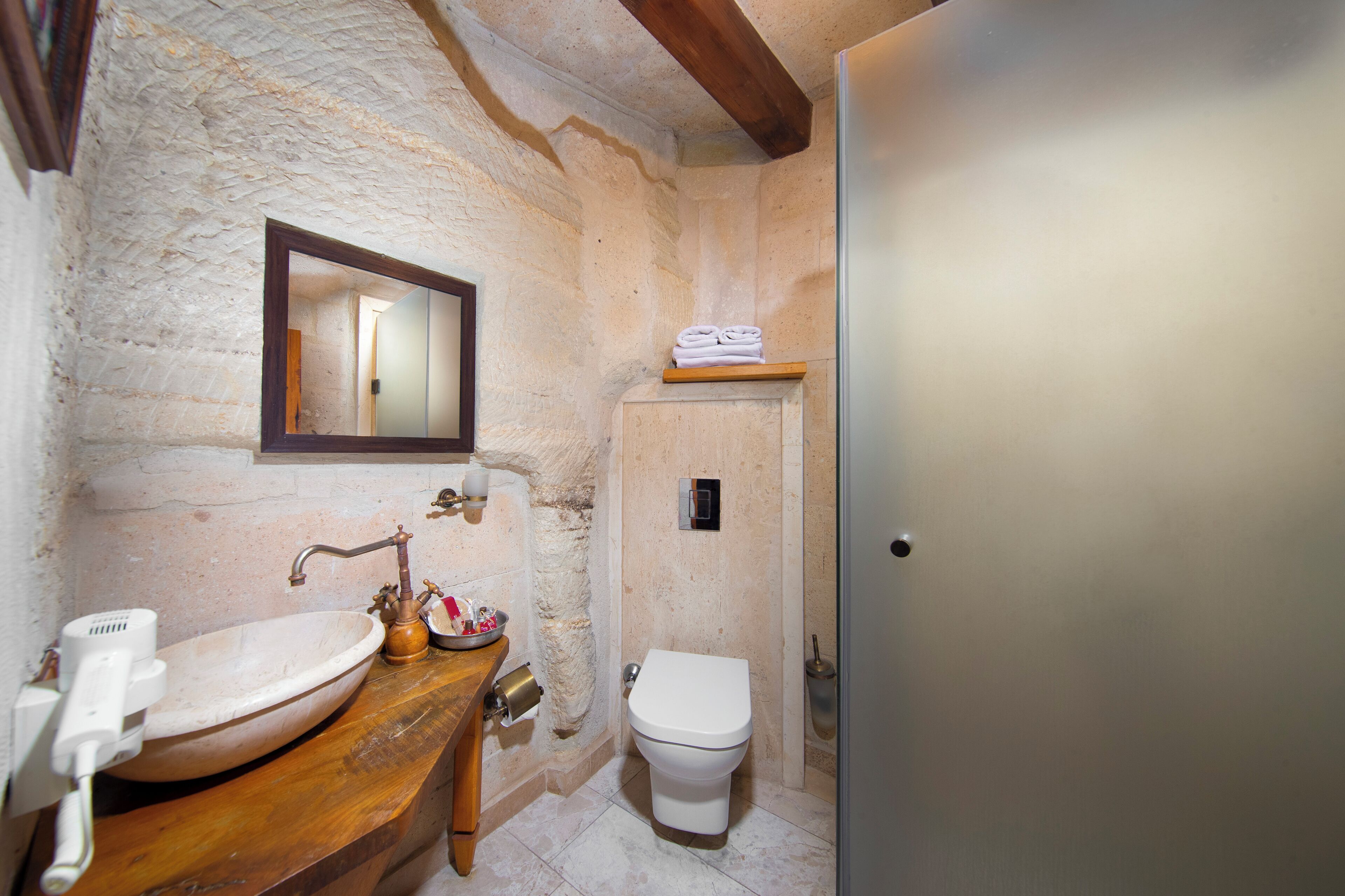 A bathroom with a stone wall, wooden vanity, a white sink, a toilet, and a mirror.
