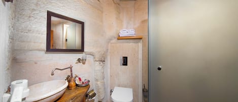 Un baño con pared de piedra, tocador de madera, lavamanos blanco, inodoro y espejo.