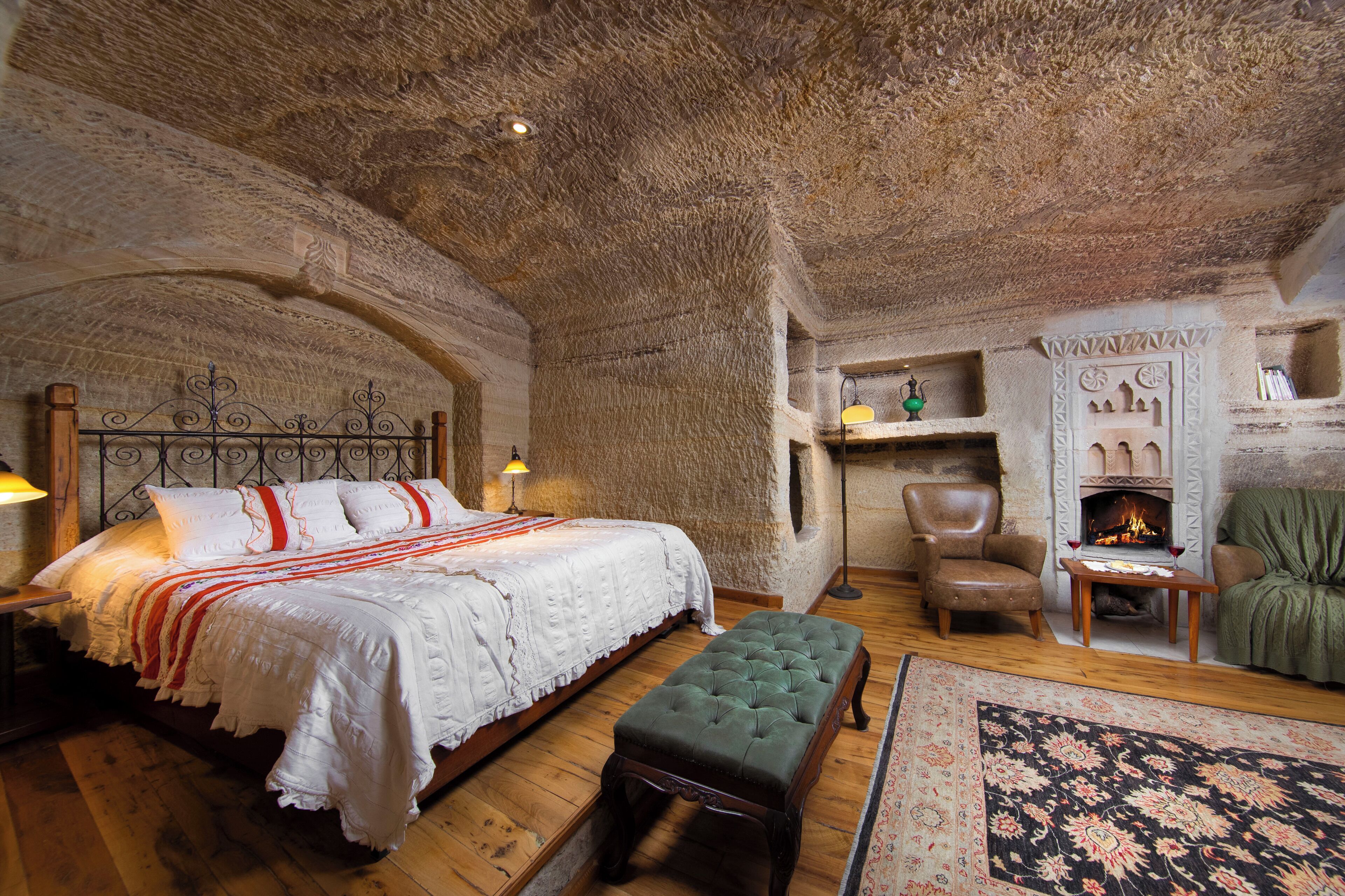 A rustic bedroom with a large bed, a green ottoman, a fireplace, and a stone ceiling.