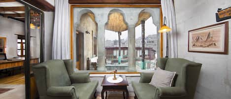 A room with two green armchairs, a wooden table, and a view through a window with arches.