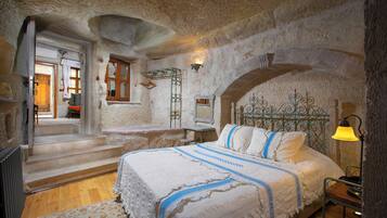 A bedroom with a stone wall, a bed with a white and blue bedspread, a nightstand with a lamp, and a staircase leading to another room.
