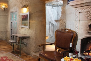 A rustic room with a stone wall, a wooden chair, a small table, a fireplace, and a shower area.