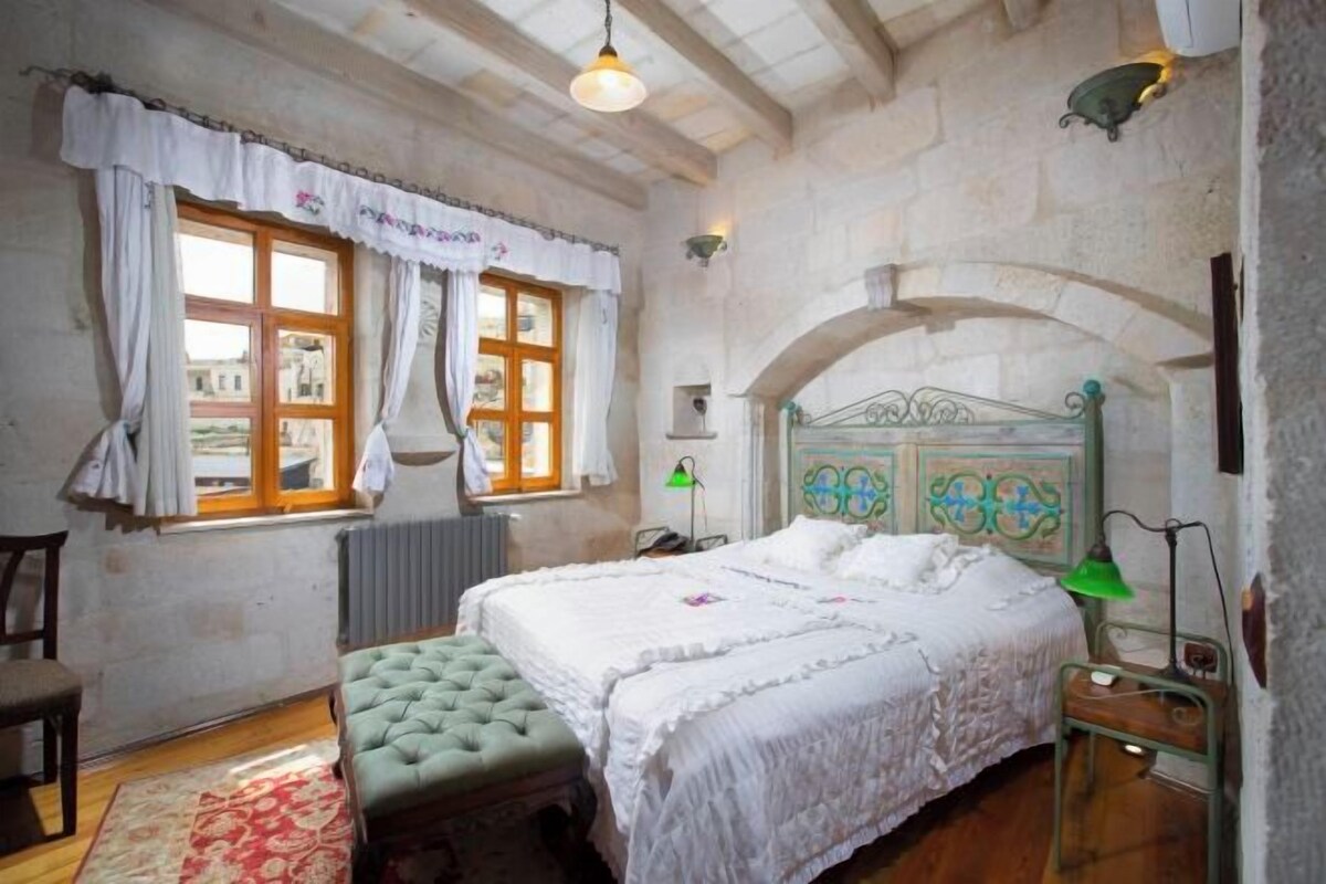 A bedroom with a large bed, a green upholstered bench, a wooden chair, and a window with curtains.
