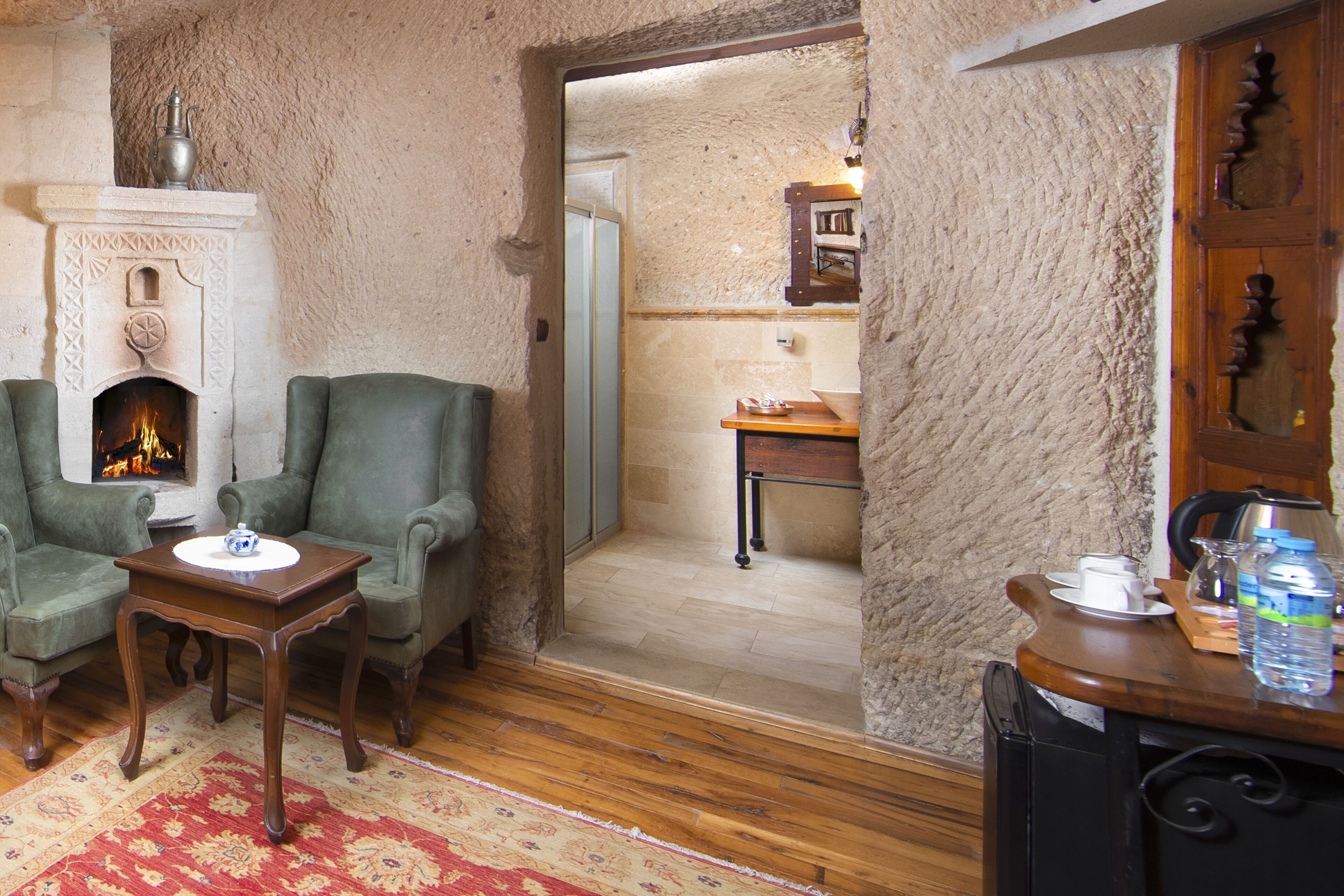 Una sala de estar acogedora con chimenea, dos sillones verdes, una mesa de centro de madera y una alfombra roja con estampado.