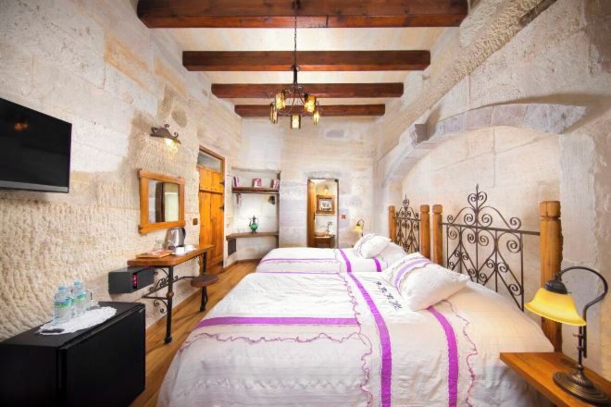 Un dormitorio rústico con paredes de piedra, vigas de madera, dos camas con ropa blanca y detalles morados, un escritorio con espejo y un televisor en la pared.