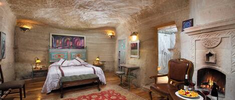 A rustic bedroom with a large bed, a fireplace, and a dining table set with fruit and wine.