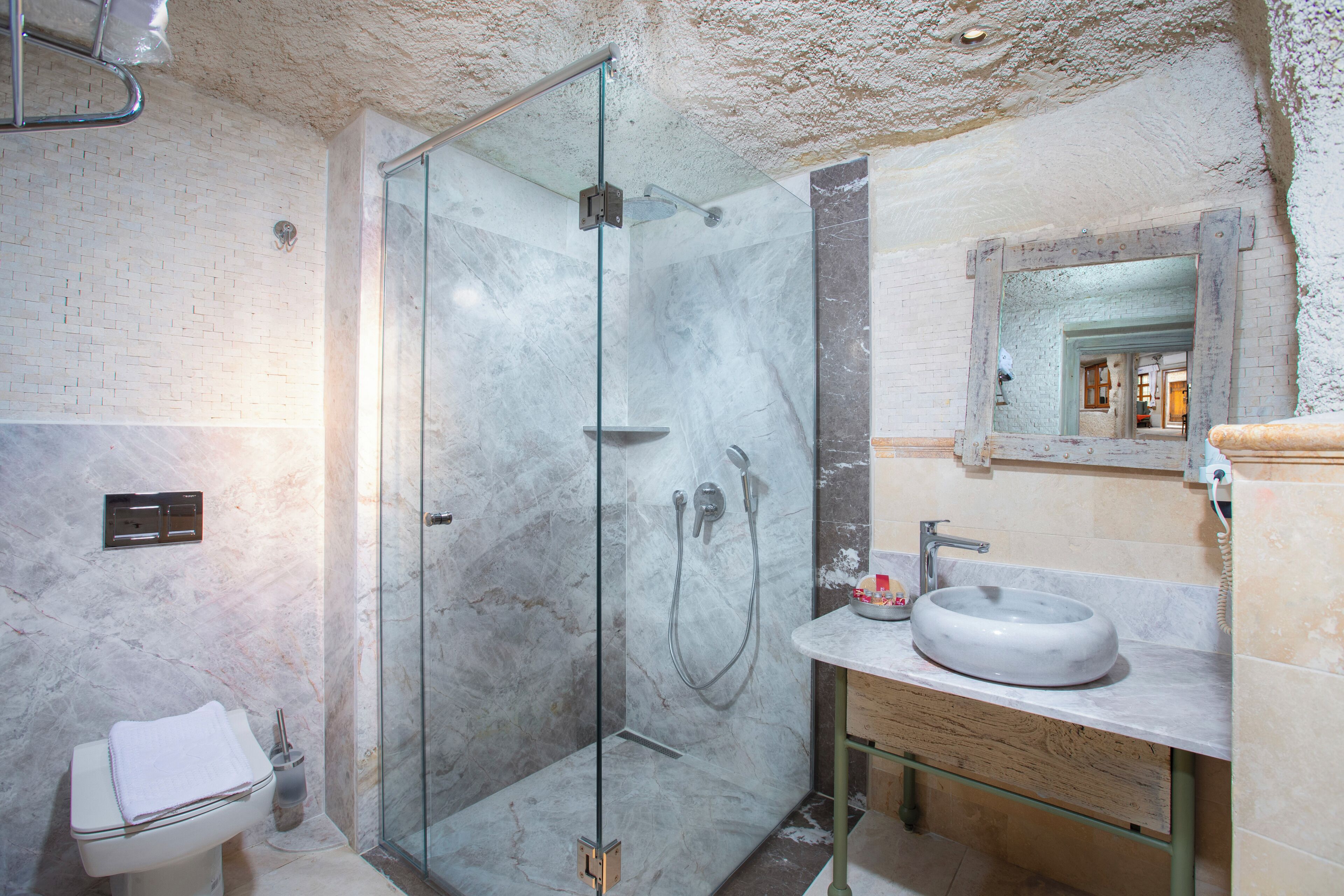 Un baño moderno con ducha envuelta en vidrio, pared con azulejos de mármol, lavamanos con encimera de mármol y tocador de madera.