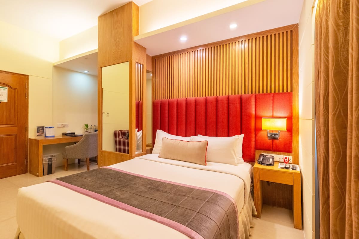 A hotel room with a large bed, a desk, a chair, a mirror, and a nightstand with a lamp.