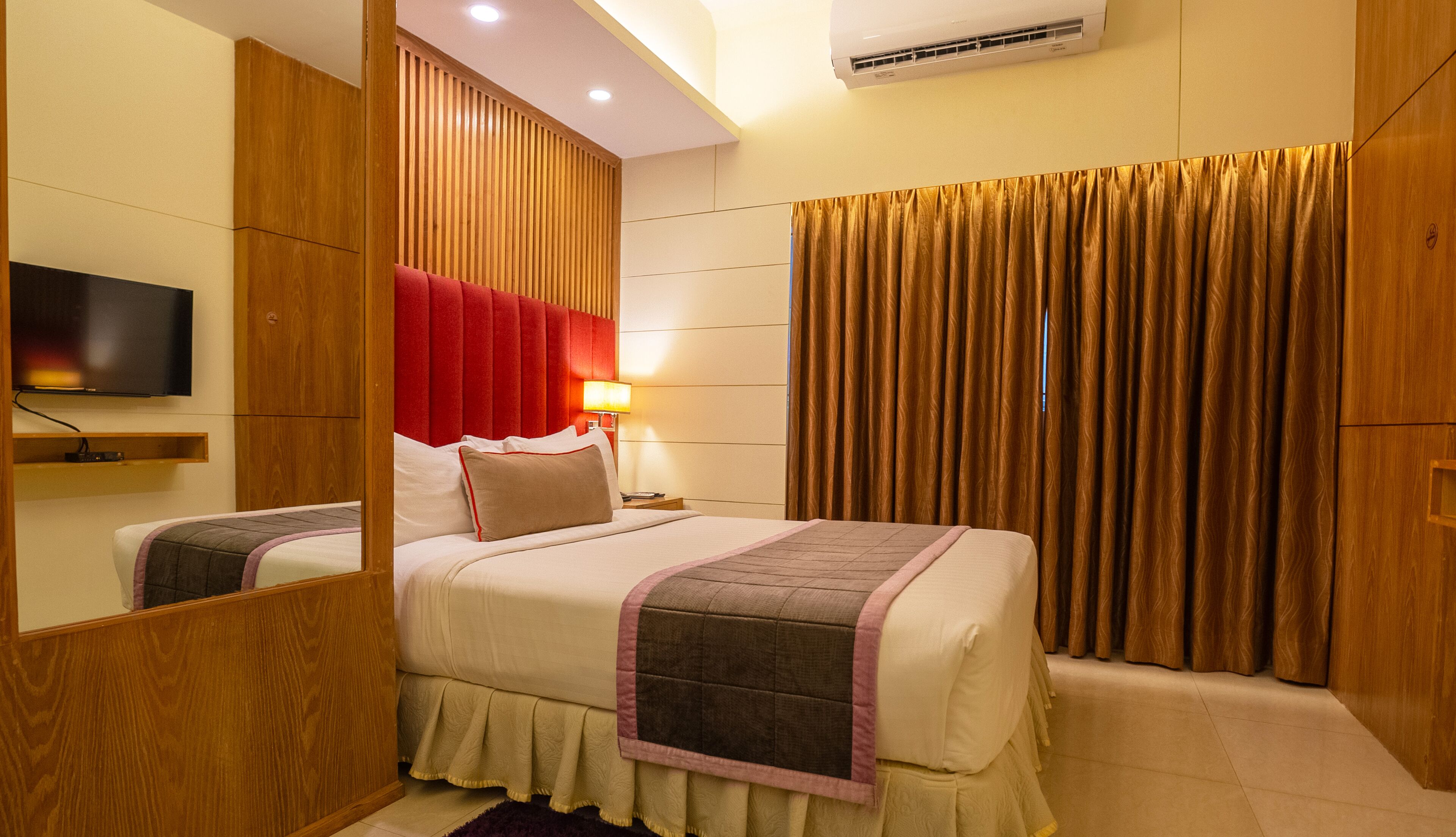 A hotel room with a bed, a TV, a bedside table, a lamp, and a large window with curtains.