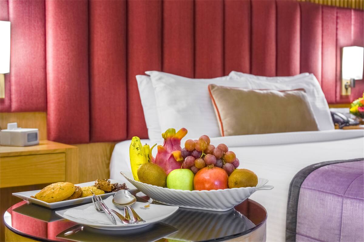 A hotel room with a bed, a bedside table with a fruit bowl, a plate with pastries, and a nightstand with a lamp.