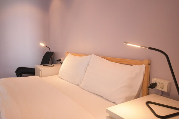 A neatly made bed with white linens, a bedside table with a lamp, and a power outlet on the wall.