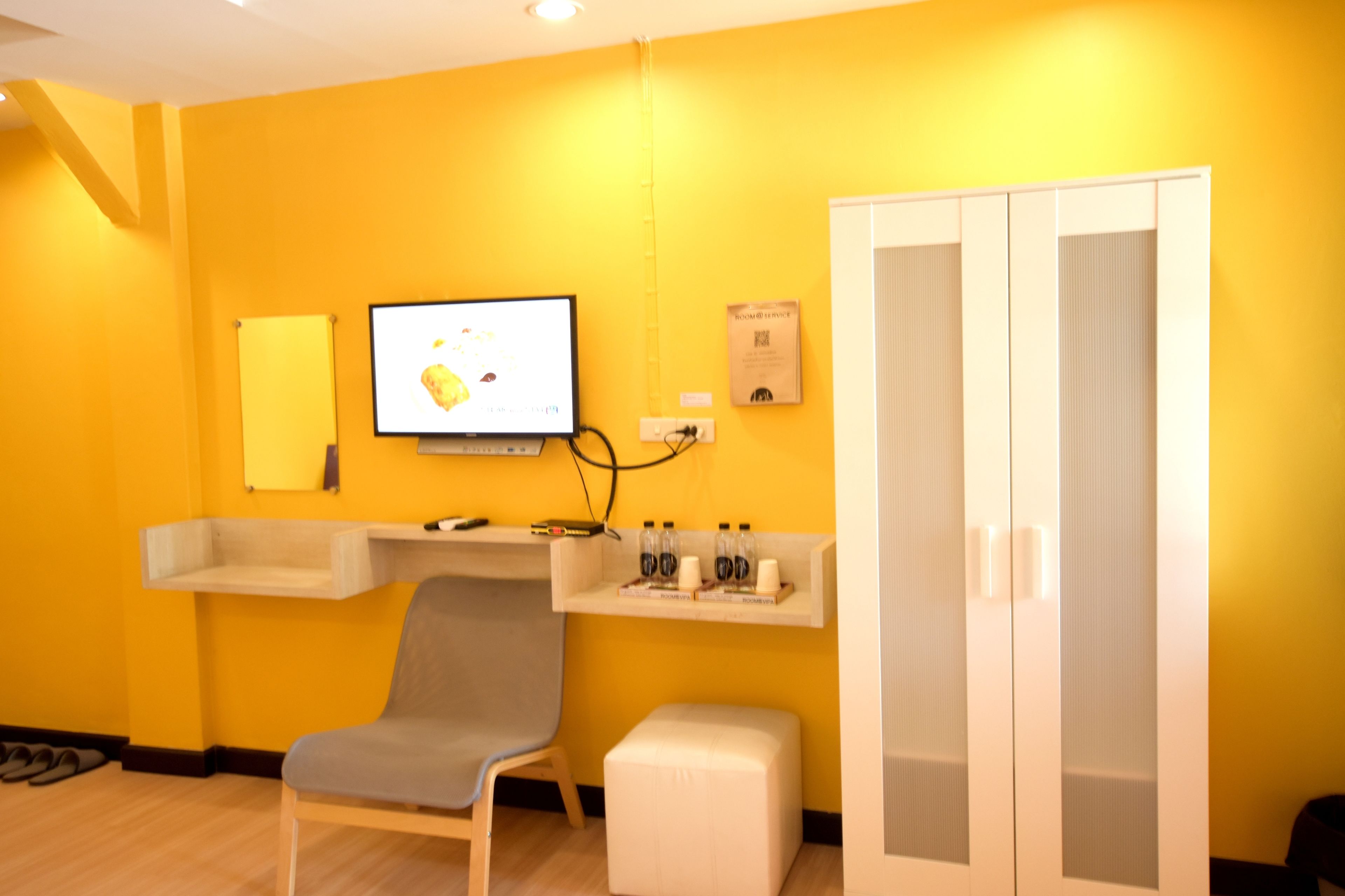 A room with a yellow wall, a TV mounted on the wall, a chair, a small table with bottles, and a wardrobe.