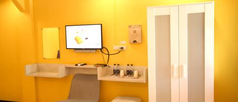 A room with a yellow wall, a TV mounted on the wall, a chair, a small table with bottles, and a wardrobe.