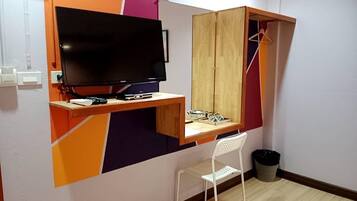 A small room with a wooden desk, a television, a mirror, a white chair, and a trash bin.