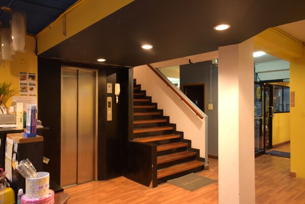 Un vestíbulo de hotel con una escalera de madera, un mostrador de recepción y una puerta de cristal que da a otra zona.