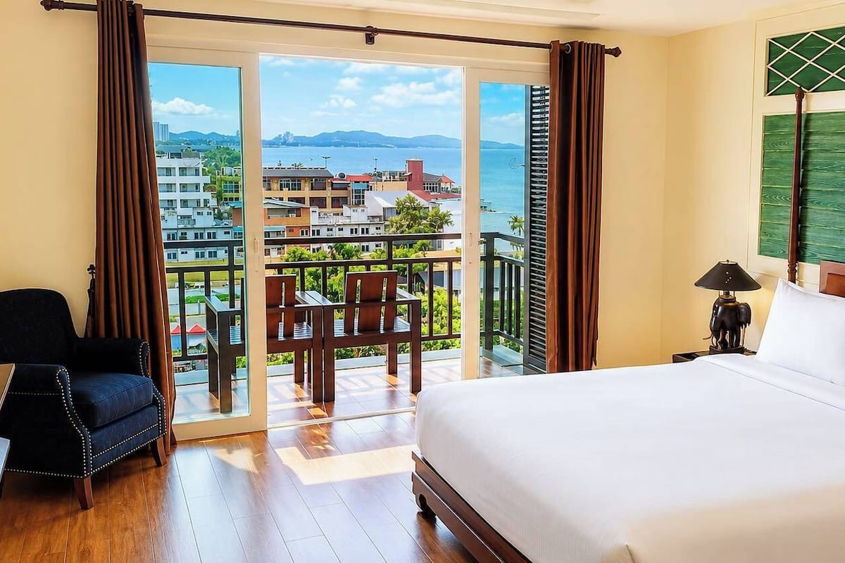 Ein Hotelzimmer mit einem großen Bett, einem Schreibtisch mit Stuhl, einem Balkon mit Blick auf die Stadt und einem dekorativen Kopfteil.