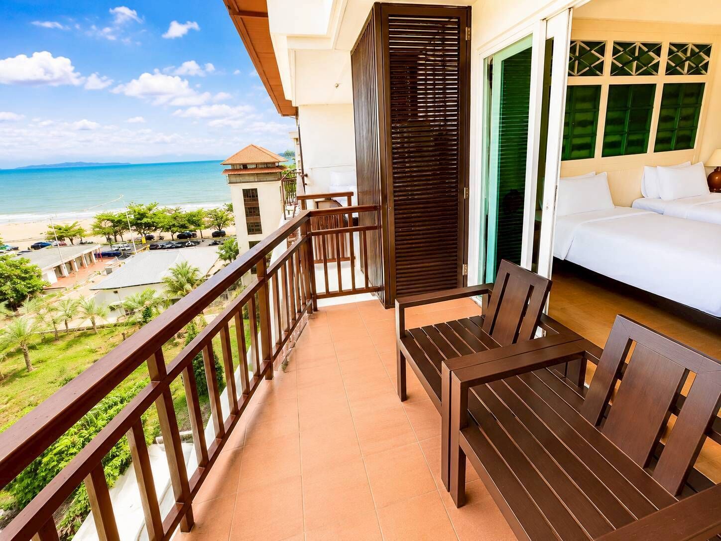 Uma varanda com mesa e cadeiras de madeira, cama com roupa de cama branca e vista para a praia e o oceano.