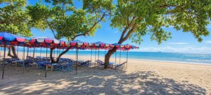 A beach with rows of sun loungers under colorful umbrellas, a clear blue sky, and the ocean in the background.