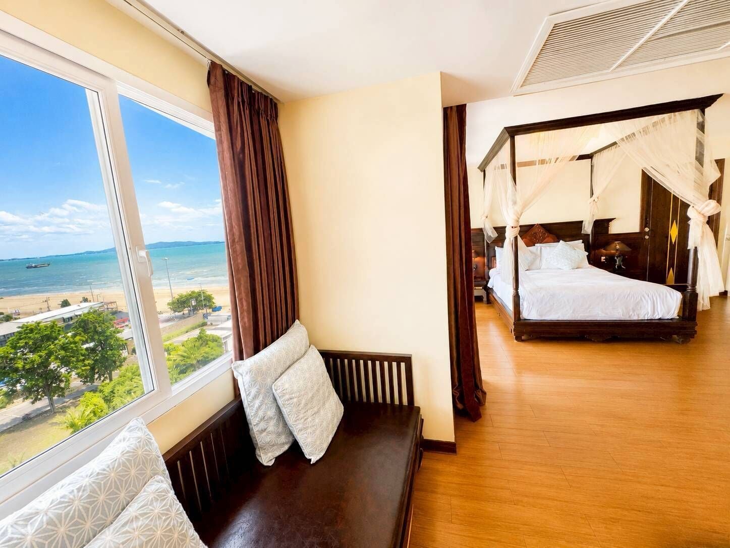 Quarto com uma grande janela com vista para uma praia, uma cama com dossel e um banco com almofadas.