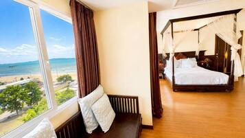 Quarto com uma grande janela com vista para uma praia, uma cama com dossel e um banco com almofadas.