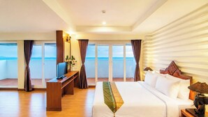 A hotel room with a large bed, a desk with a TV, a balcony with curtains, and a view of the sea.