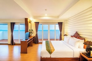 A hotel room with a large bed, a desk with a TV, a balcony with curtains, and a view of the sea.