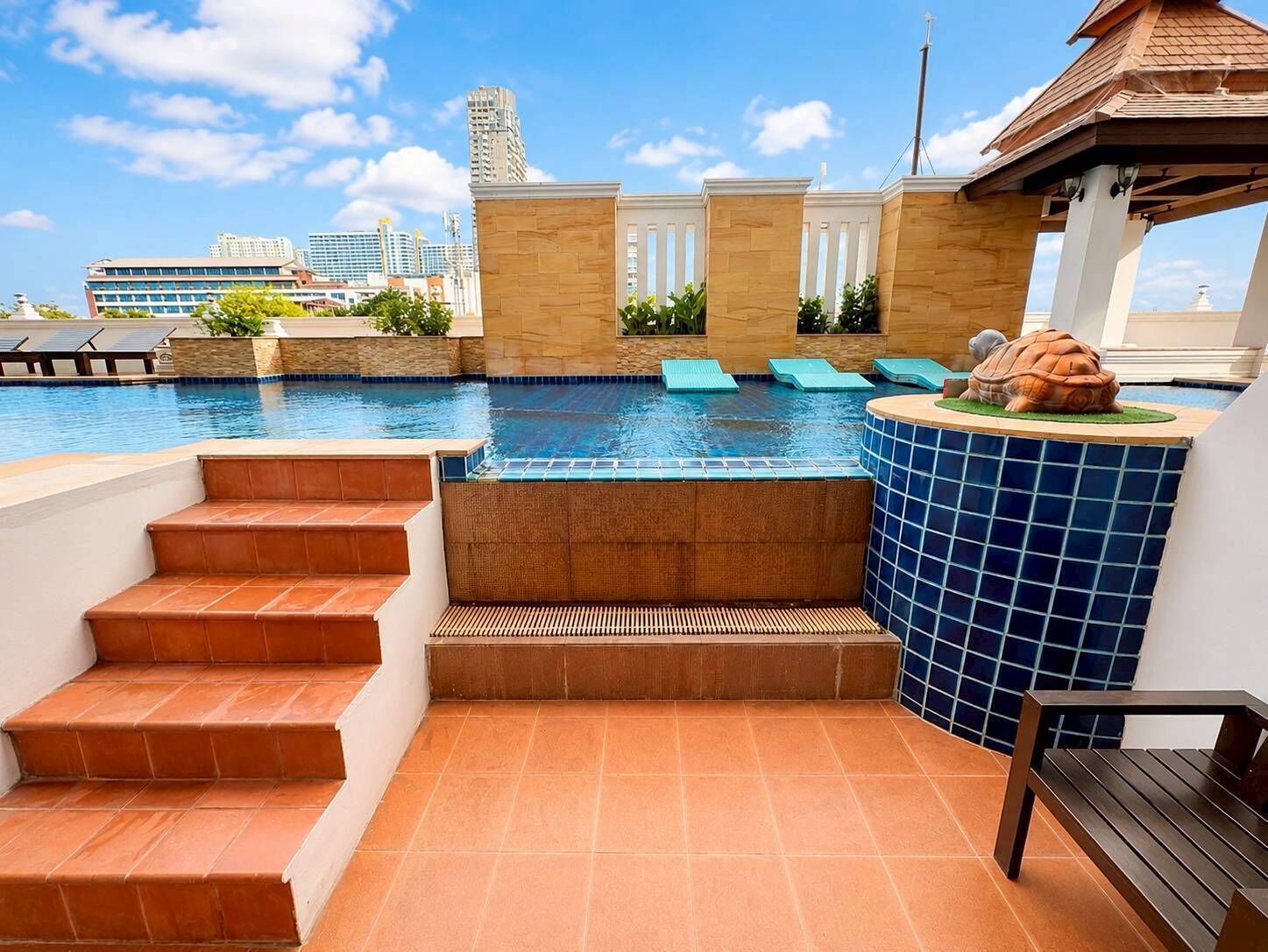 A rooftop pool with steps leading down to a hot tub area, surrounded by a tiled deck and a view of city buildings.