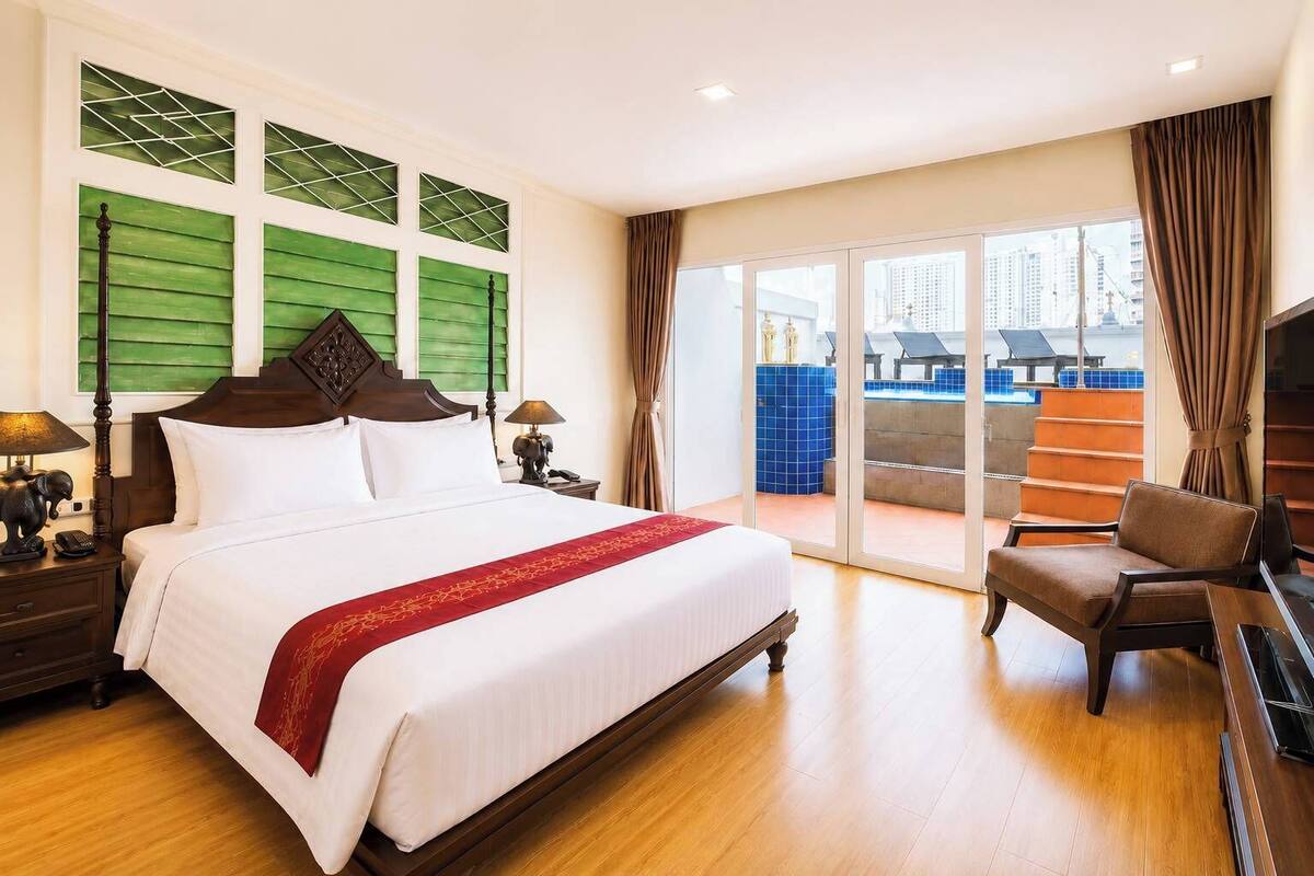 Een hotelkamer met een groot bed, een stoel, een bureau en uitzicht op een zwembad en gebouwen.