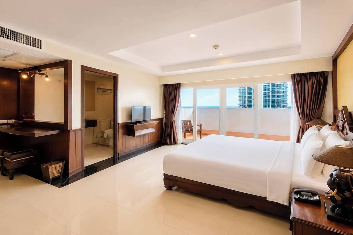 Ein Hotelzimmer mit einem großen Bett, einem Schreibtisch mit Fernseher, einem Balkon mit Blick auf Gebäude und einem Badezimmer mit Dusche und Toilette.