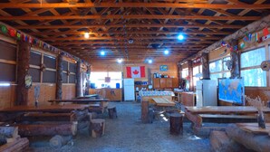 A rustic wooden interior with a high ceiling, wooden beams, and a Canadian flag. There are wooden benches and tables, a kitchen area, and a large window. - Mountain View Cabins (Golden)