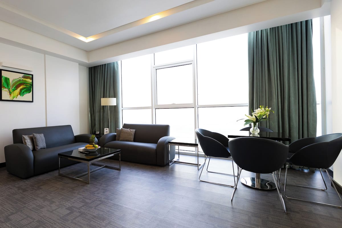 A modern hotel room with a dark grey sofa, a coffee table, and a dining area with black chairs. There is a large window with curtains, a painting on the wall, and a bed in the background.