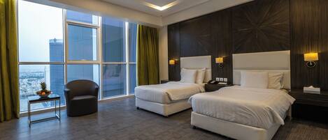 A hotel room with two beds, a chair, a small table with a fruit bowl, and a view of the city.