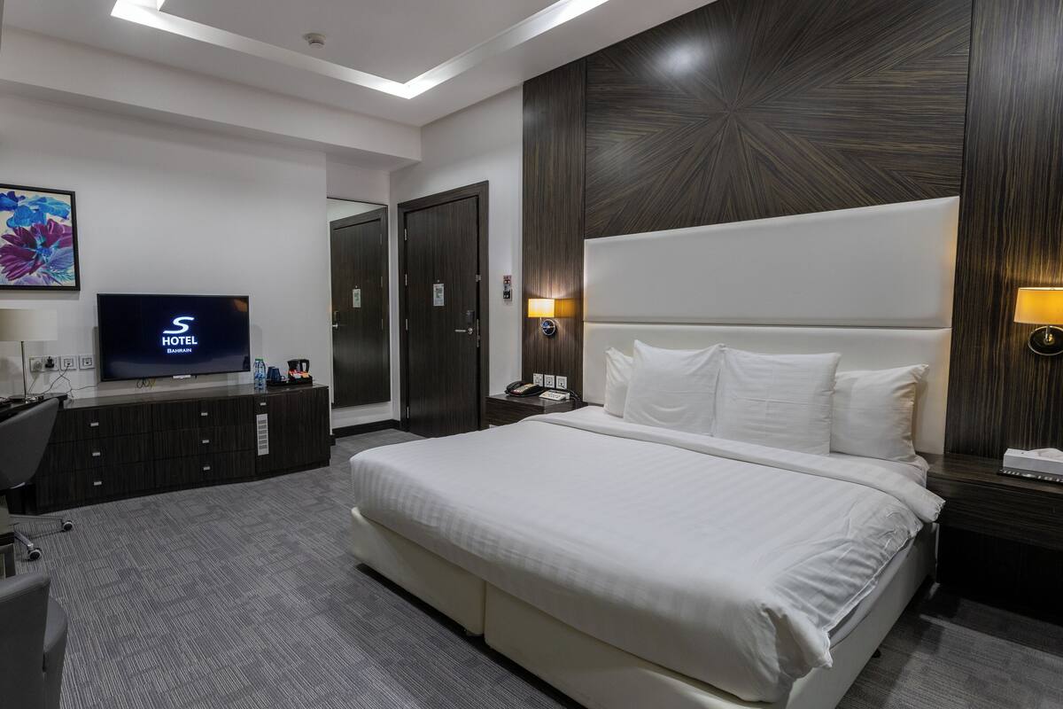 A hotel room with a large bed, a desk with a computer, a TV displaying 'S HOTEL BAHRAIN', a mirror, and a wall-mounted lamp.