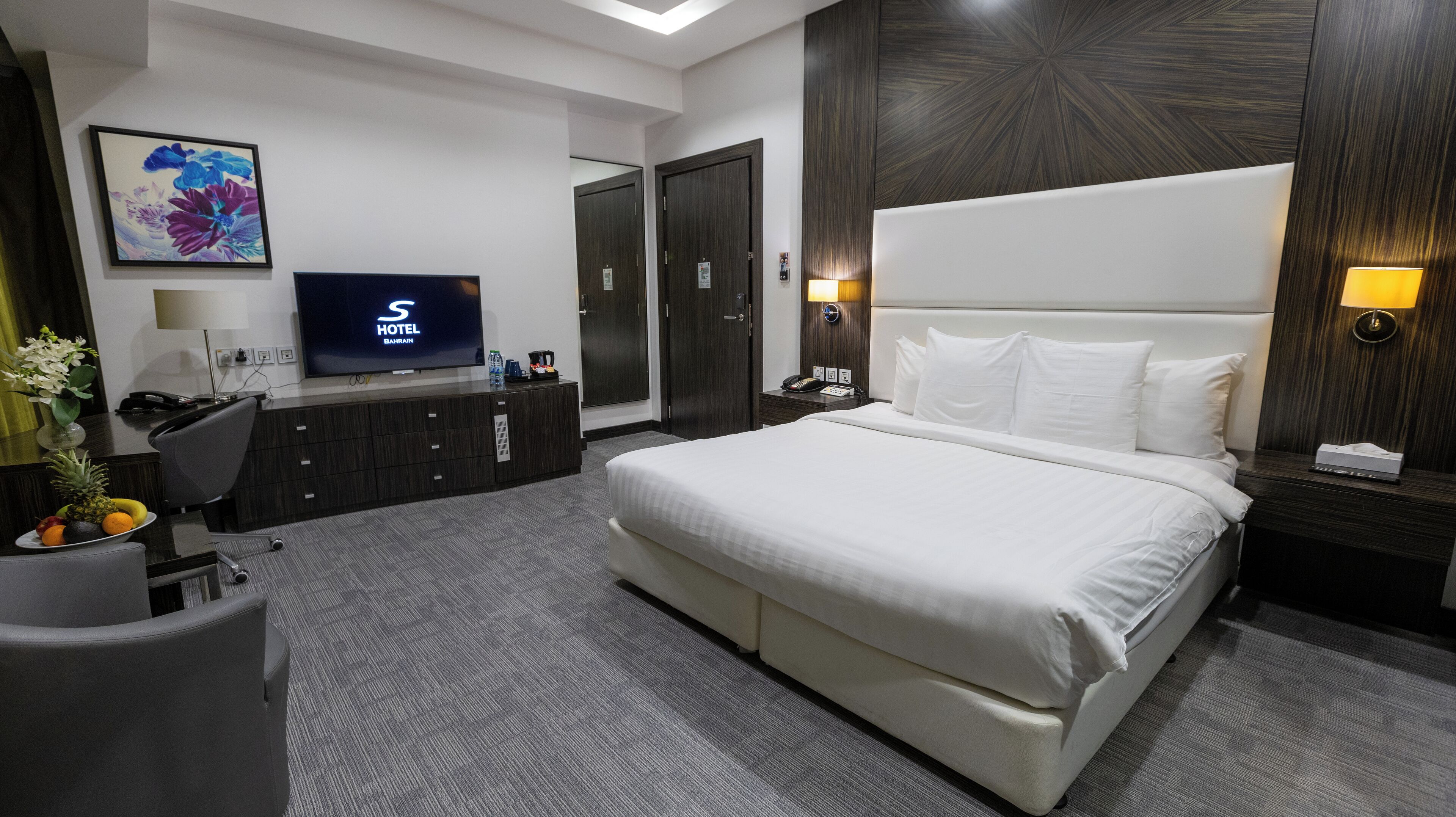 A hotel room with a bed, a desk with a TV, a desk lamp, a chair, a plant, and a wall-mounted artwork.